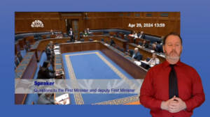 An ISL interpreter in front of a screenshot of Questions to the Executive Office in the Plenary Chamber on Monday 29 April 2024.