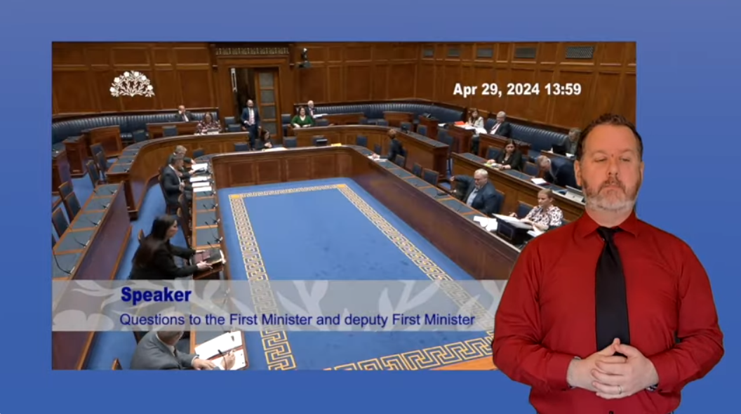 An ISL interpreter in front of a screenshot of Questions to the Executive Office in the Plenary Chamber on Monday 29 April 2024.