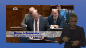 Still shot from the Assembly Chamber showing Gordon Lyons, Minister for Communities, answering a question during Question Time. In the foreground, an Irish Sign Language interpreter is shown providing interpretation.
