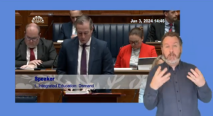 Minister for Education Paul Givan, stands in the Assembly Chamber to answer a question during Question Time on Monday 3 June 2024. In the foreground we see an ISL interpreter translating what the Minister is saying for the hard of hearing.