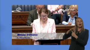 Finance Minister Caoimhe Archibald speaking in the Assembly Chamber. In the foreground we see a BSL interpreter translating what the Minister is saying for the hard of hearing.