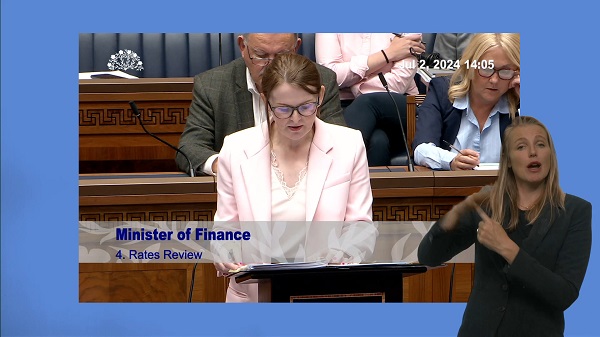 Finance Minister Caoimhe Archibald speaking in the Assembly Chamber. In the foreground we see a BSL interpreter translating what the Minister is saying for the hard of hearing.