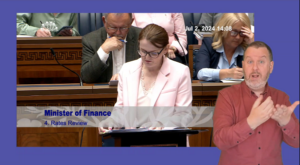 Finance Minister Caoimhe Archibald speaking in the Assembly Chamber. In the foreground we see a ISL interpreter translating what the Minister is saying for the hard of hearing.