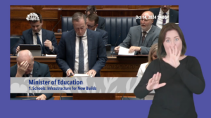 Paul Givan, Minister of Education, answering Members questions in the Assembly Chamber on Monday 14 October 2024. In the foreground, an ISL interpreter interprets Mr Givan's answers.