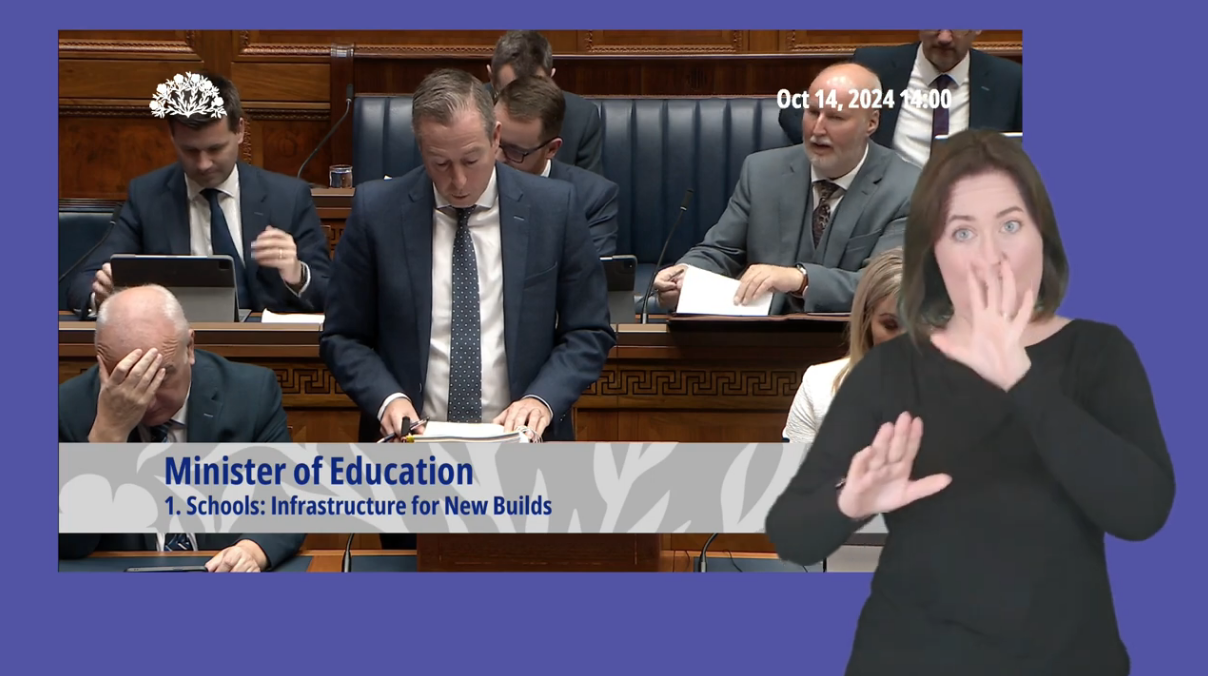 Paul Givan, Minister of Education, answering Members questions in the Assembly Chamber on Monday 14 October 2024. In the foreground, an ISL interpreter interprets Mr Givan's answers.